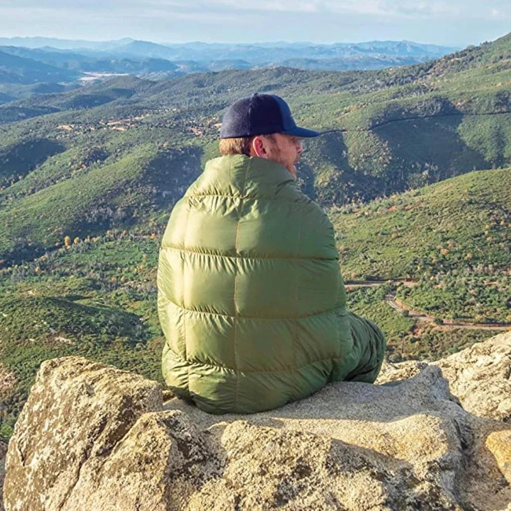 Man sitting on a mountain ledge wearing the Adventure Cozy Puff Ultralight Blanket, looking at the valley landscape