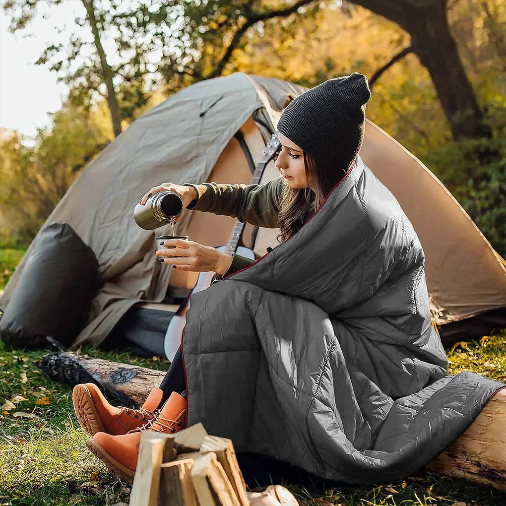 Woman sitting outside a tent pouring coffee, wrapped in the Adventure Cozy Puff Ultralight Blanket, at a campsite